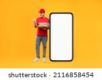 Food Delivery Application. Courier Guy Near Big Smartphone Holding Pizza Boxes And Using Mobile Phone Standing On Yellow Studio Background, Wearing Red Uniform. Mockup, Full Length