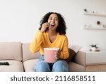 Lovely young black woman eating popcorn from bucket while watching TV on couch at home. Cheery millennial African American female enjoying interesting movie or program on television