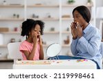 Small photo of Female speech-language pathologist young black woman exercising with adorable little girl with bushy hair, working on word sonds, kid looking at mirror and touching her mouth, clinic interior