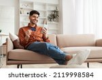 Small photo of Fast Online Shopping. Smiling young Arab guy holding debit credit card in hand and using cell phone, making financial transaction sitting on the couch at home in living room, free copy space