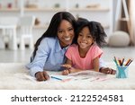 Small photo of Portrait of cheerful loving african american mom and little girl with bushy hair daughter coloring together, laying on carpet at kids room, painting, embracing and smiling at camera, copy space