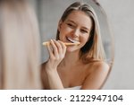 Oral hygiene, healthy teeth and care. Young woman brushing teeth with toothbrush and looking in mirror in bathroom interior in the morning, closeup, empty space