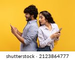 Small photo of Shocked indian woman spying on her smiling boyfriend who using smartphone, chatting or scrolling social media news feed. Young couple standing back to back over yellow studio background