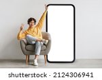 Small photo of Joyful guy sittiwng on chair near big empty smartphone screen, listening to music in wireless headphones over grey wall. Musical application. Mockup