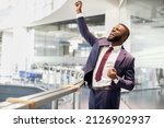 Cheerful african american young businessman celebrating success, handsome black manager in suit and tie raising hands up and screaming, office building interior, panorama with copy space