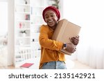 Small photo of Ecstatic young african american muslim woman in casual embracing box with something and smiling, home interior, making order on Internet with delivery, copy space. Delivery service, shipment concept