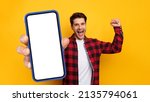 Small photo of Yes, Big Luck. Excited Guy Holding Big Smart Phone In Hand Showing Empty White Screen Shaking Clenched Fist Raising Hand Up, Cheerful Man Celebrating Win, Looking At Camera, Mock Up Collage Banner