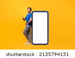 Small photo of Cool Mobile Offer. Excited casual guy leaning on big smartphone with blank empty white display, showing thumb up gesture, recommending great new app or website, mock up, full body length, yellow wall