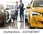Small photo of Customer middle eastern guy standing among brand new cars, having conversation with male sales assistant, looking at catalogue in dealer hands, checking auto features, full length, blurred background