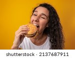 Small photo of Gluttony Concept. Closeup Portrait Of Funny Hungry Lady Biting Burger Eating Junk Food On Yellow Orange Studio Background, Enjoying Big Hamburger. Unhealthy Nutrition And Binge Eating Habit