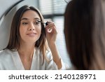 Beautiful Millennial Lady Doing Daily Makeup Near Mirror In Bathroom, Attractive Young Woman Applying Mascara On Eyelashes, Holding Brush And Smiling To Her Reflection, Selective Focus, Closeup