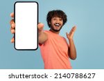 Emotional handsome curly young indian man in stylish pink t-shirt showing brand new cell phone with white empty screen and gesturing, sharing exciting online deal, blue background, mockup
