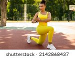 Small photo of Sport And Healthy Lifestyle. Fit black woman doing curtsy lunges with twist, exercising outdoors on basketball stadium. Portrait of confident lady training in wireless earbuds, full body length