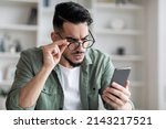 Eyesight Problems Concept. Young Arab Man In Eyeglasses Looking At Smartphone Screen And Frowning, Millennial Guy Trying To Read Message, Suffering From Astigmatism And Bad Vision, Closeup Shot