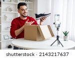 Fashion Blog. Young Arab Male Influencer Recording Video Content At Home, Smiling Middle Eastern Guy Unpacking Box With New Shoes In Front Of Camera, Using Smartphone On Tripod, Copy Space