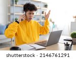 Small photo of Confused Discontented Asian Guy Talking On Cell Phone Using Pc Having Problem With Computer Or Internet Connection At Home. Displeased Male Customer Calling To Hotline Service And Complaining