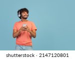 Happy millennial eastern guy in pink t-shirt using brand new wireless stereo headset and smartphone, listening to music over blue studio background, looking at copy space and smiling, panorama