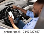 Over the shoulder view of happy black male driver holding smartphone with white blank screen, using mobile app for navigation, mock up. Closeup of cell phone with empty display, copy space template