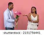 Small photo of Portrait of African American man giving pretty unhappy bored lady bunch of ugly cheap flowers. Young boyfriend holding present for his ungrateful girlfriend isolated on pink studio background