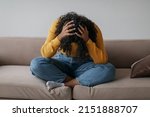 Depressed young black woman sitting cross legged on sofa with head in her hands at home. African American female patient feeling stressed, suffering from PTSD or having psychological problem