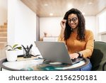 Small photo of People And Technology. Portrait of young black woman wearing eyeglasses using pc sitting at table on beanbag chair in living room, typing on keyboard. Cheerful lady browsing internet, free copy space