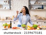 Small photo of Young woman eating salad at table with organic vegetables, enjoying healthy diet, standing in light kitchen interior. Lady cooked veggie meal at home. Weight loss concept