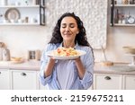 Happy lady tasting homemade spaghetti while having lunch, enjoying the smell with eyes closed, sitting in kitchen interior. Woman cooking and eating tasty food at home