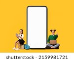 Small photo of Wow, Great Offer. Excited Multiethnic School Girls Sitting On Floor And Pile Stack Of Books Near Big Giant Cell Screen, Using Pc Laptop And Tablet On Yellow Orange Studio Background, Showing Thumbs Up