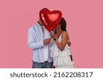 St. Valentines Day Concept. Shy African American couple in love kissing, holding and hiding behind red heart shaped balloon, covering their faces for privacy, isolated on pink studio background wall