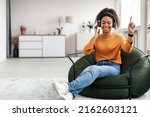 Small photo of Overjoyed black woman wearing wireless headphones dancing sitting on beanbag chair, happy attractive young female listening to popular music, moving to favorite song, full body length, free copy space