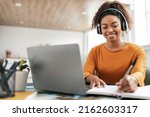 Smiling young black woman in wireless headphones sitting at desk table working on laptop and writing letter in paper notebook, taking notes watching weninar, holding pen in hand, free copy space