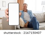 Smiling senior man showing big blank smartphone with white screen at camera while sitting on couch at home, happy elderly male recommending mobile website or app, creative collage, mockup