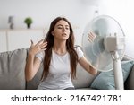 Small photo of Sad young european woman suffers from unbearably too hot weather, catches cold air from fan in living room interior. Home without air conditioning, lady tired from summer heat, waves arms to cool down