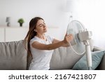 Small photo of Glad young european woman suffers from heat sits on sofa catches cold air, turns fan to herself, enjoys cool down in living room interior. Summer weather, overheating without air conditioning at home