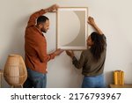 Small photo of Black Husband And Wife Hanging Picture In Frame On Wall At Home. Married Couple Decorating Room With Poster Together. Interior Design, Art And Decoration Concept