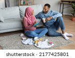 Small photo of Pregnant black muslim couple making checklist of necessities while getting ready for childbirth, happy islamic spouses awaiting baby, relaxing on floor in living room with child clothes around