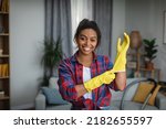 Smiling young african american woman puts on rubber gloves and ready to homework in living room interior. Housewife has fun and many household chores, domestic work and professional cleaning service