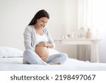 Small photo of Happy pregnant woman sitting on bed in bedroom at home, hugging her big tummy. Happiness and feeling joyful as emotional well affect to baby neurological and psychological development, empty space
