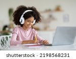 Small photo of Cheerful african american schooler cute preteen girl with bushy hair sitting at table in front of laptop, using wireless headset, taking notes, doing homework at home, copy space