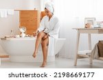 Pretty Woman Shaving Legs Using Safety Razor Sitting In Modern Bathroom At Home, Wearing White Bathrobe. Depilation And Bodycare, Hair Removal Cosmetics Concept