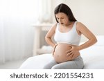 Small photo of Braxton Hicks contractions. Young pregnant woman in pain suffering from abdominal ache at home, sitting on bed, touching her big tummy, having cramp, panorama with free space