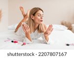 Gorgeous middle aged woman doing manicure, applying nail polish and blowing on nails while lying on bed at home. Mature lady making self-care procedure indoors. Domestic spa salon