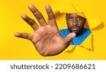 Small photo of Serious african american man reaching through hole in torn yellow paper, trying to grab something. Black male grasping, trying to start fight, feeling annoyed, panorama