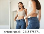 Small photo of Slimming Concept. Excited Slim Woman Wearing Jeans After Great Weight Loss Comparing Size Posing Standing Near Mirror At Home. Selective Focus, Cropped Shot