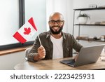 Small photo of Happy latin man sitting with flag of Canada and using laptop computer, sitting at desk at home. Modern online foreign education, emigration and citizenship, working abroad concept