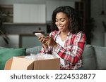 Happy pretty black young woman client in casual unpacks parcel, takes photo of purchases on smartphone in living room interior. Fashion blog and online shopping, gadget for reviews on buy and delivery