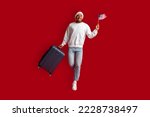 Excited handsome millennial african american guy in casual and Santa hat holding American flag, carrying suitcase, jumping over red studio background, travelling to the US on xmas holidays, copy space