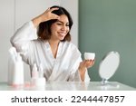 Skin care concept. Beautiful lady using new cosmetic at home, attractive middle eastern young woman in white silk robe sitting in front of mirror and applying moisturising face cream, copy space