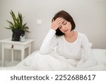 Tired unhappy young beautiful brunette woman waking up with headache in the morning, sick lady sitting in bed at home, touching her head, suffering from hangover or migraine, copy space