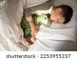 Small photo of Happy cute tired little boy lies on white comfort bed with closed eyes, hugs toy dinosaur, sleeping in bedroom interior, top view, free space, close up. Health care, childhood, rest, relax at night